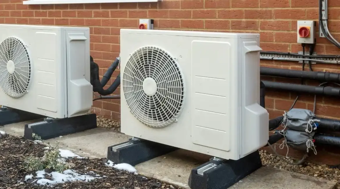 Two white outdoor air conditioning condenser units on concrete pads beside a brick wall with exposed pipes